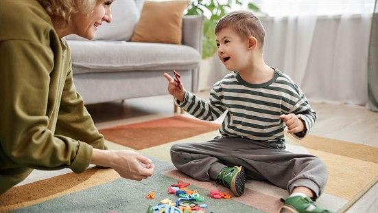Illustrasjonsfoto av et barn med Downs syndrom som leker sammen med sin mor på stuegulvet. Barnet er glad og leker med bokstavformede brikker.