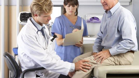 Fotoet viser en yngre mannlig lege som undersøker det venstre kneet til en eldre mann. Mannen sitter på  en undersøkelsesbenk ikledd skjorte og bukse. Imellom og bak legen og pasienten står en helsesekretær, som skriver notater.