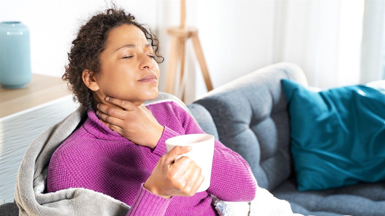 Illustrasjonsbilde viser en mørk kvinne som sitter i sofaen. Hun har en kopp varm te i hånden, og holder den andre hånden sin over halsen. Man ser hun har vondt i halsen. 