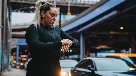 Illustrasjonsbilde av en kvinne som er på treningstur i en travel bygate og ser på klokken sin.  