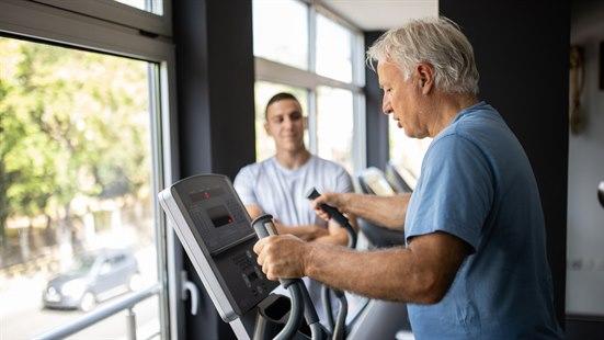 Illustrasjonsfotoet viser en eldre man som trener på et apparat. I bakgrunnen ser man en person som veileder.