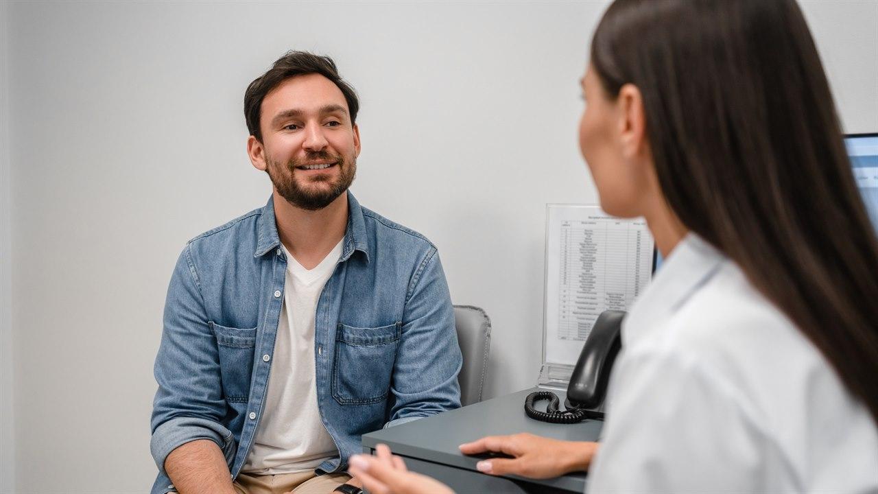 Illustrasjonsbilde viser en ung mann som er på legebesøk. Den kvinnelige legen lytter og forklarer.