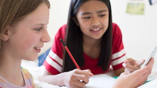 Illustrasjonsfoto av to jenter i 12-årsalderen som sitter og gjør lekser og ser på mobiltelefonen.
