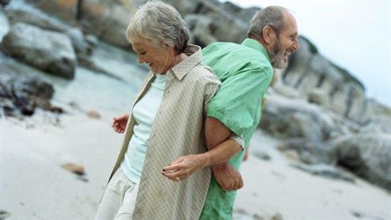 Illustrasjonsfoto som viser et eldre par, kvinne og mann som står rygg mot rygg på en strand og smiler.