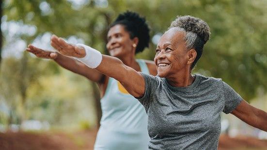 Illustrasjonsfoto av to glade og mørkhudet kvinner som trener sammen i en park. De står med den ene hånden rett frem og den andre hånden rett bak.