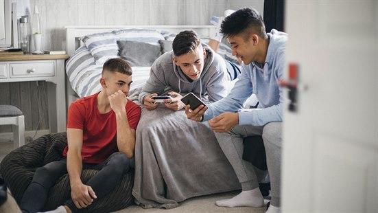 Illustrasjonsfoto av tre tenåringsgutter, en i sakkosekk ved sengen, en avslappet i sengen, og den tredje viser begeistret noe på mobilen til de to andre.