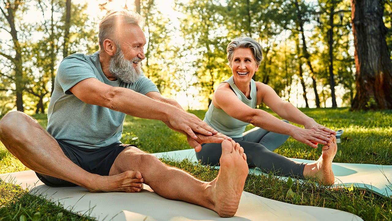 Illustrasjonsfoto av en mann og kvinne med grått hår som sitter på en yogamatte i parken og tøyer baksiden av lårene.