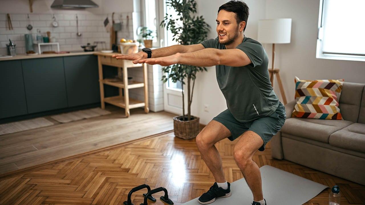 Illustrasjonsfoto av en ung mann med mørkt hår og grønne treningsklær som utfører en squat-øvelse i stuen. I bakgrunnen kan man se kjøkkenbenken.