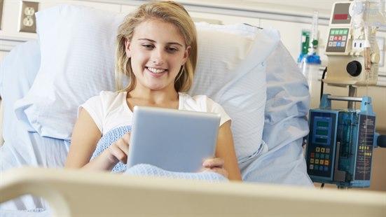 Illustrasjonsfoto av en ung kvinne som ligger i sykehussengen og ser på et nettbrett.