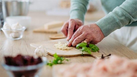 Illustrasjonsfoto: Colourbox. Nærbilde av hender som kutter hvitløk på et skjærebrett. Genserermene er grågrønne. 