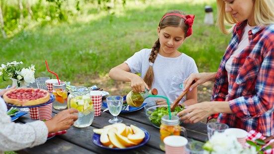 Illustrasjonsfoto av jente og kvinne som spiser mat utendørs en sommerdag fra et bord fullt av salat og annen mat.