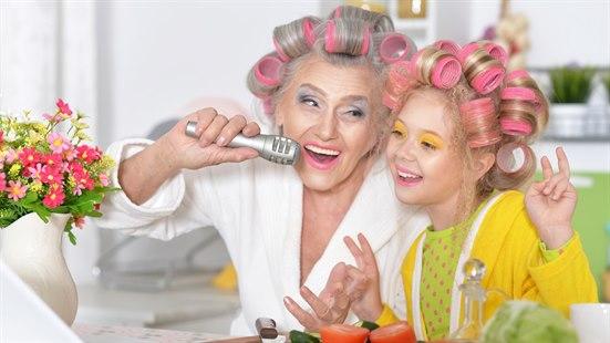 Illustrasjonsfoto: Bestemor og barnebarn med hårruller, morgenkåpe og fargerik sminke synge ri en mikrofon ved kjøkkenbordet.