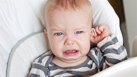 Fotoet viser overkroppen og ansiktet til en gutt på ca. 10 måneder foran hvit bakgrunn. Han er ikledd en body med grå og hvite striper. Han gråter og tar seg til det venstre øret sitt med sin venstre hånd. 