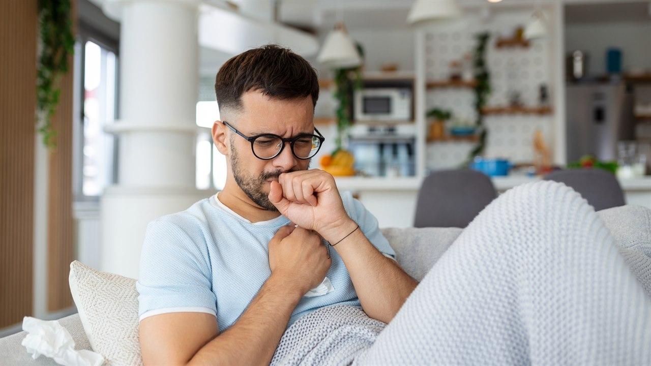 Illustrasjonsbilde viser en mann som ligger på sofaen og ser syk ut. Han holder en hånd på brystet og en hånd foran munnen mens han hoster.