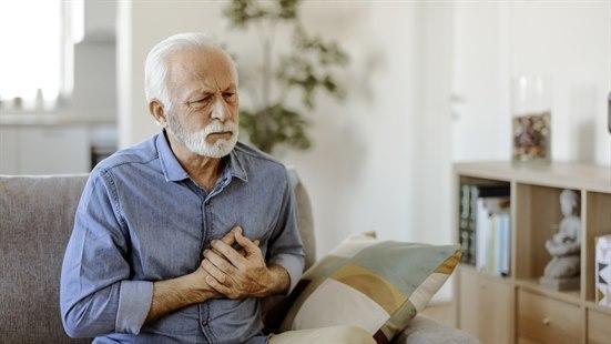 Illustrasjonsfotoet viser en eldre mann som sitter i en sofa og holder hendene foran hjertet. Han har brystsmerter og ser bekymret ut.