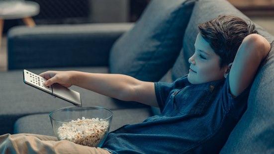 Illustrasjonsfoto av en mørkhåret gutt sitter i sofaen med fjernkontroll i hånden og en skål popcorn på fanget. Gutten sitter i en grå sofa og har blå skjorte og beige bukser.