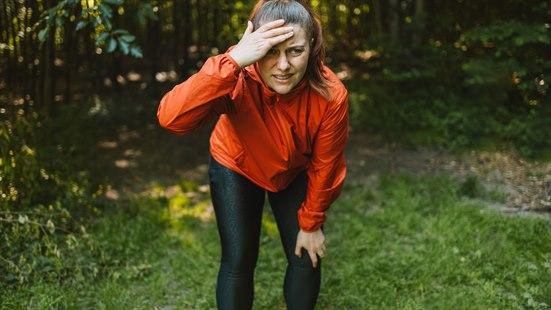 Illustrasjonsfoto: Kvinne i løpeklær holder seg for pannen med den ene hånden og på kneet med den andre. Bakgrunnen viser skog og grønt gress.