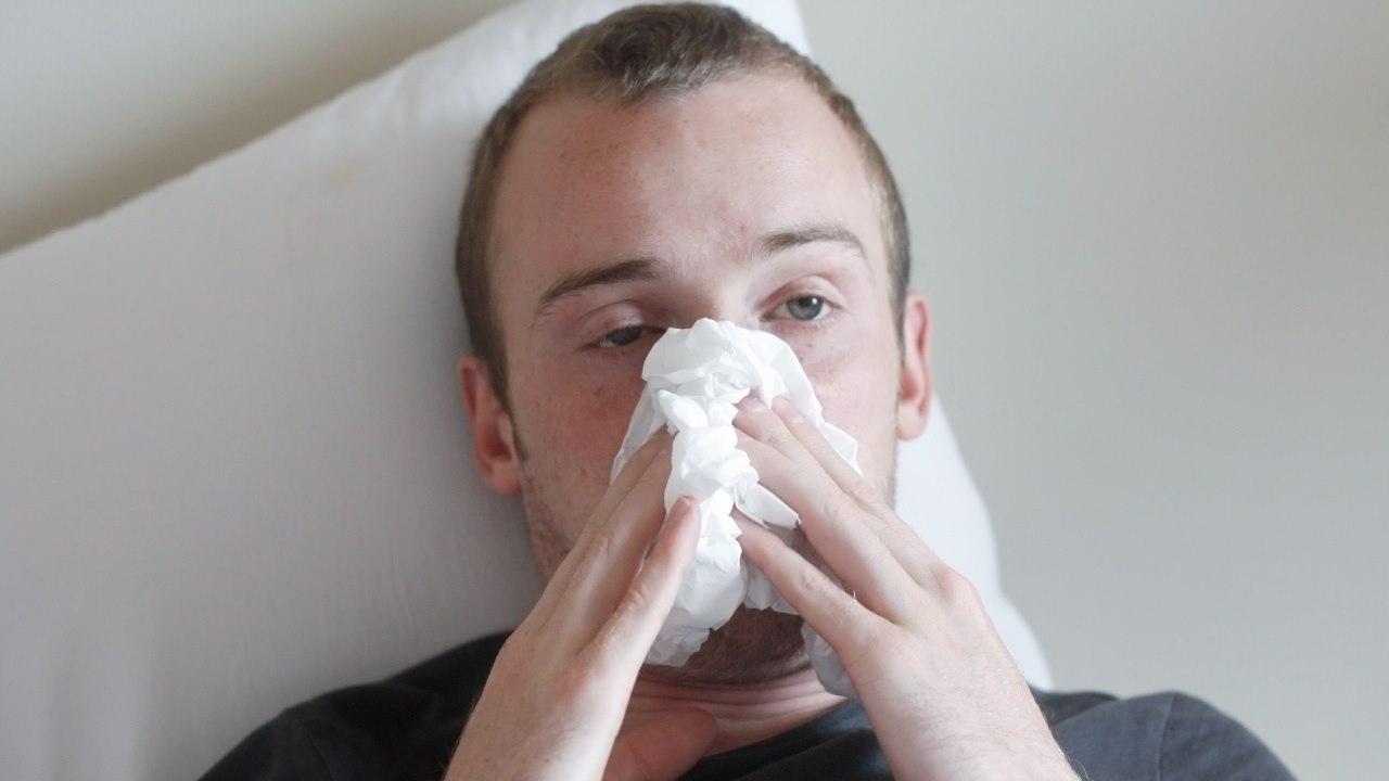 Illustrasjonsfotoet viser en forkjølet mann som ligger i sengen og holder et snytepapir foran nesen. Han ser syk og sliten ut.