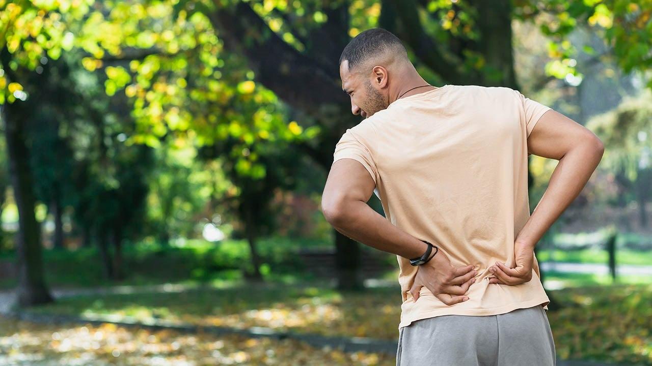 Illustrasjonsfoto av en ung mørkhudet mann i lyse treningsklær. Han holder seg på korsryggen, noe som tyder på smerte.