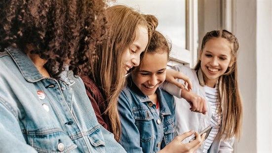 Illustrasjonsfoto av fire jenter lener seg mot en husvegg. En av dem viser noe på mobilen og ler, mens resten ser nysgjerrige og engasjerte ut.