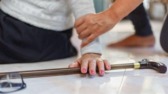 Illustrasjonsfotoet viser en eldre dame som har falt. Fotoet viser en stokk, armen og deler av kroppen til damen. I tillegg ses en annen hjelpende hånd