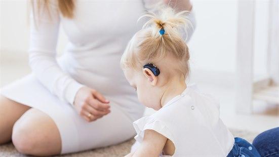 Illustrasjonsfoto: Lita jente med høreapparat sitter med ryggen til. En dame i hvit kjole sitter foran henne,.