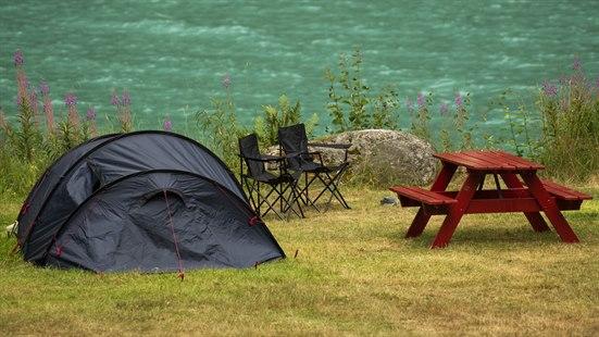 Illustrasjonsfoto av et telt som er oppslått ved et vann. To campingstoler står ved siden av. 