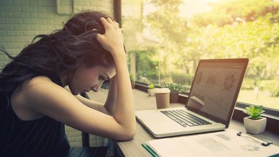 Ung kvinne sitter på en kafe og arbeider. Hun ser oppgitt ut og holder begge hendene på hode.