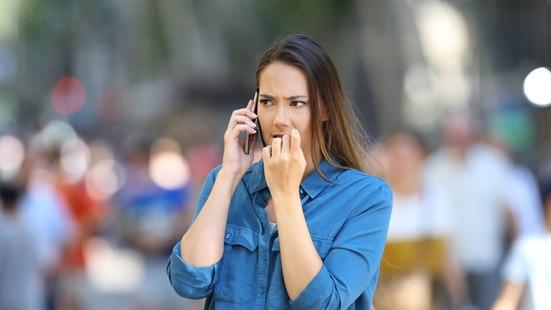 Illustrasjonsfoto: En kvinne med blå skjorte står i en folkemengde. Hun holder en mobiltelefon mot øret, biter negler og ser engstelig ut.