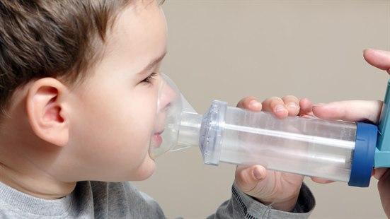 Fotoet viser høyre siden av ansiktet til en cirka fem år gammel gutt som holder en inhalasjonskolbe med ansiktsmaske med sin venstre hånd mot ansiktet. På høyre kanten av bildet vises det inhalatoren på enden av kolben som utløses av en hjelper.