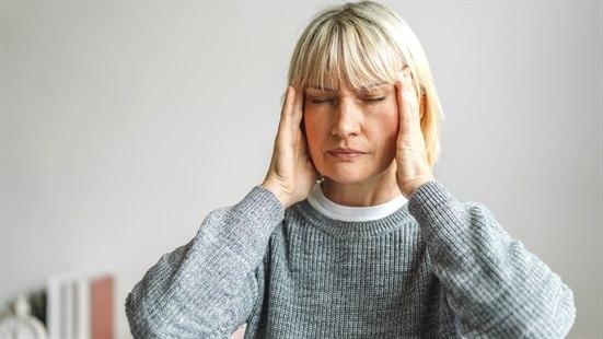 Illustrasjonsfoto: Middelaldrende, blond kvinne med grå genser som masserer tinningene med lukkede øyne.
