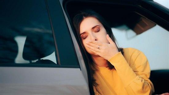 En kvinne sitter i passasjersetet og vendt ut mot veien. Hun lener hodet mot bilen samtidig som hun holder seg for munnen. Hun ser uvel og kvalm ut.
