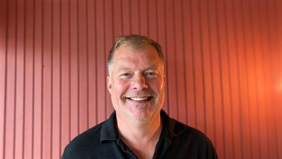 Smilende mann i svart skjorte står foran rød vegg. Utsnitt fra skuldrene og opp.