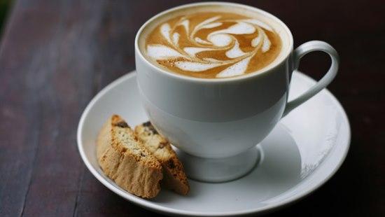 Hvit kaffekopp med mønster i kaffeskum. På et tefat sammen med biscotti