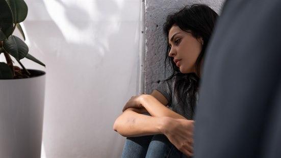 Ung kvinne med langt mørkt hår hviler hodet mot en hvit murvegg. Holder hendene rundt knærne og ser trist ut.