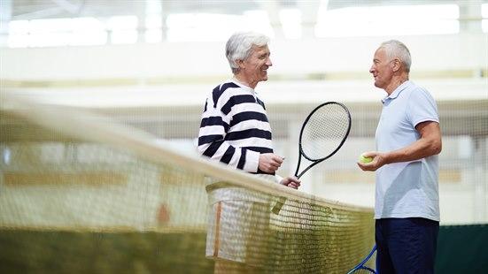 To eldre menn som står ved et tennisnett med tennisracketer.
