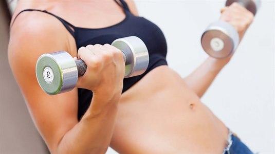 En kvinne løfter vekter på treningsstudioet. Hun holder en manual i hver hånd og utfører Bicepscurls med manualer. Hun har god kroppsholdning.