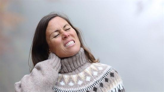 Illustrasjonsfoto: Kvinne i grå ullgenser holder seg til nakken, ser ut som hun har det vondt.