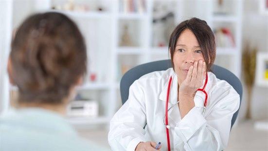 Illustrasjonsfoto: Kvinne i hvit frakk med rødt stetoskop rundt halsen sitter på en stol og lytter. Foran henne ser vi bakhodet til en annen kvinne.