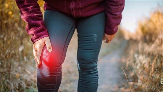 Illustrasjonsfoto av en eldre kvinne ute på en grusvei i sola, ikledd treningsklær. Utsnittet viser henne forfra, fra magen til ankelen. Hun holder sitt høyre kne med sin høyre hånd. Området rundt kneet er farget rødt.