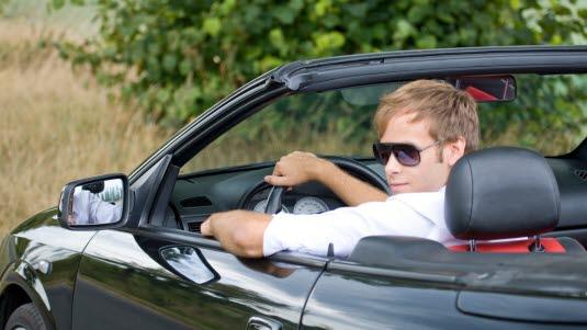 Fotoet viser en ung mann ikledd hvit skjorte som sitter i en sort kabriolet. Fotoet er tatt fra venstre og bak. Mannen snur hodet mot kameraet.