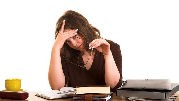 En dame sitter ved sin kontorpult og holder brillene sine i den ene hånden mens hun holder seg for pannen