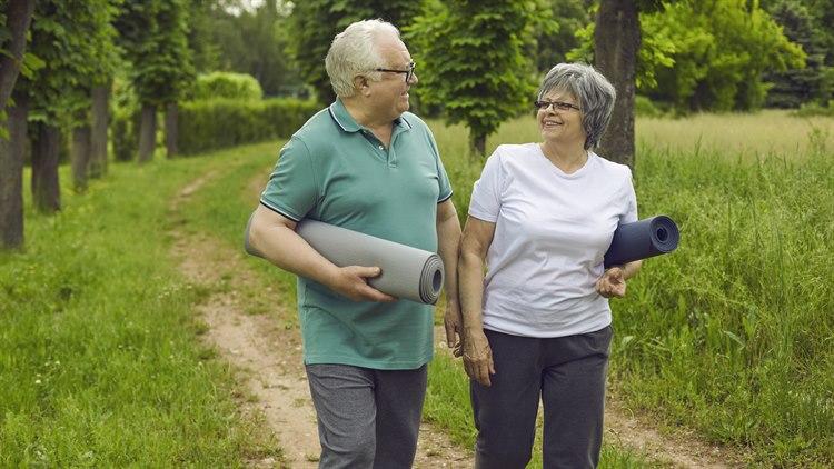 Illustrasjonsfoto av et eldre, smilende par som går på en sti med hver sin yogamatte under arma.