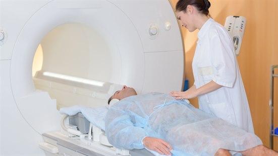 Illustrasjonsfoto: Sykepleier i hvit arbeidsdrakt står ved siden av en pasient som er på vei inn i en MR-skanner. Pasienten har en blå sykehusskjorte på seg.