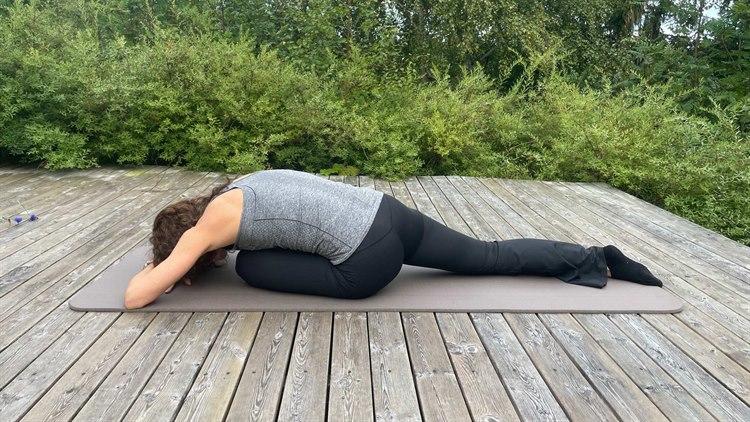 Dame som ligger fremover på yogamatte utendørs, med busker og trær i bakgrunnen. Den ene foten er strekt ut bakover, den andre er bøyd foran henne. 