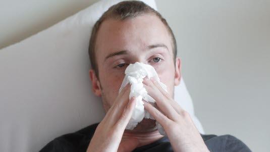 Illustrasjonsfotoet viser en forkjølet mann som ligger i sengen og holder et snytepapir foran nesen. Han ser syk og sliten ut.