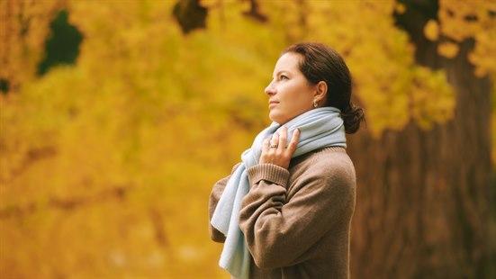 En kvinne står utendørs - det er høst og høstfarger på trærne.