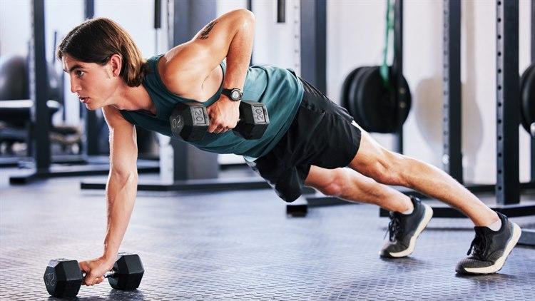 Illustrasjonsfoto: Mann på treningssenter i grønn singlet og sorte shorts gjør armhevinger med en frivekt i hver hånd.