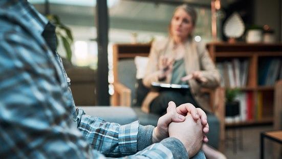 Illustrasjonsfotoet viser nærbilde av en pasient som sitter i en sofa og holder hender. I bakgrunnen ser vi en psykolog som har samtale med pasient.