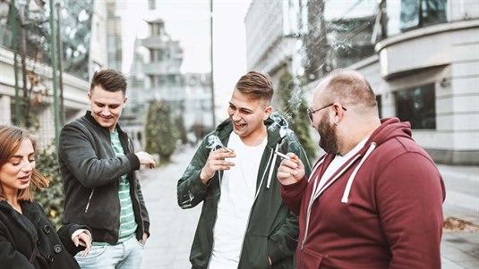 Fire personer står på gaten og røyker sigaretter. De holder hver sin sigarett i hånden og blåser ut røyk mens de snakker med hverandre.